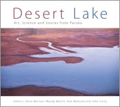 The cover image of Desert Lake, features an arial view of a dark blue lake with a river feeding into it in the middle of deep red desert.