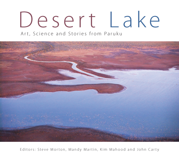 The cover image of Desert Lake, features an arial view of a dark blue lake with a river feeding into it in the middle of deep red desert.